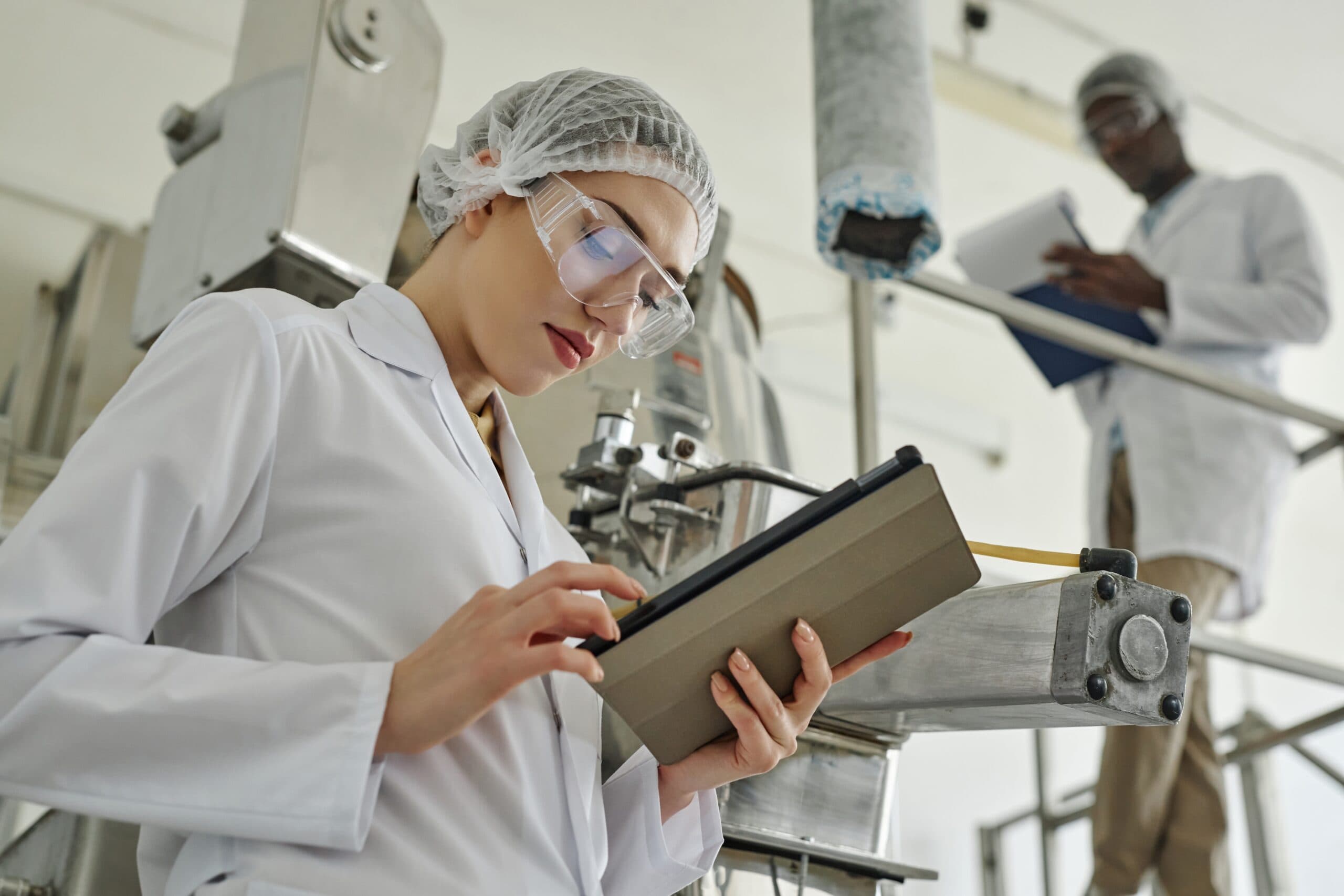 A specialist in a lab coat and safety glasses reviewing a tablet beside processing equipment